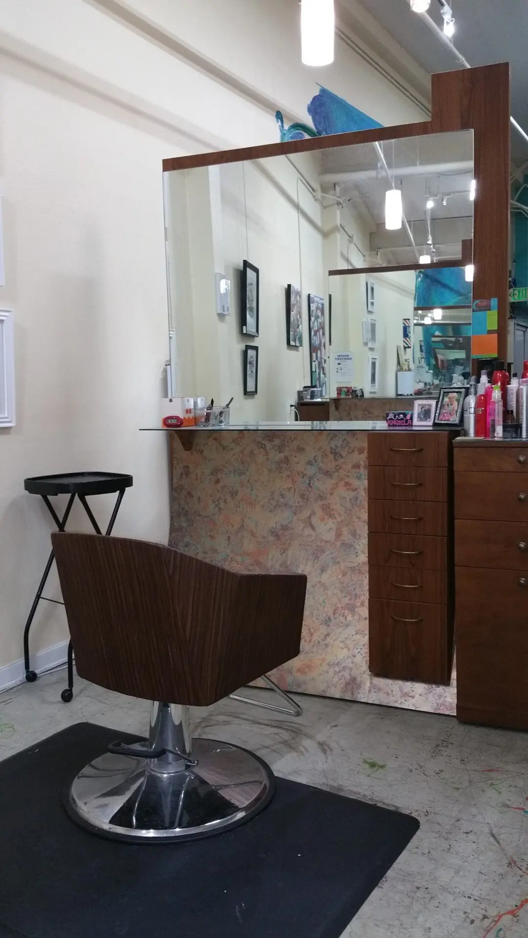 A salon station with a large mirror, a wooden chair with a metal base, and a counter holding various hair products and picture frames. The station includes a set of drawers and a side cart. The background shows an art-adorned wall and other salon stations.