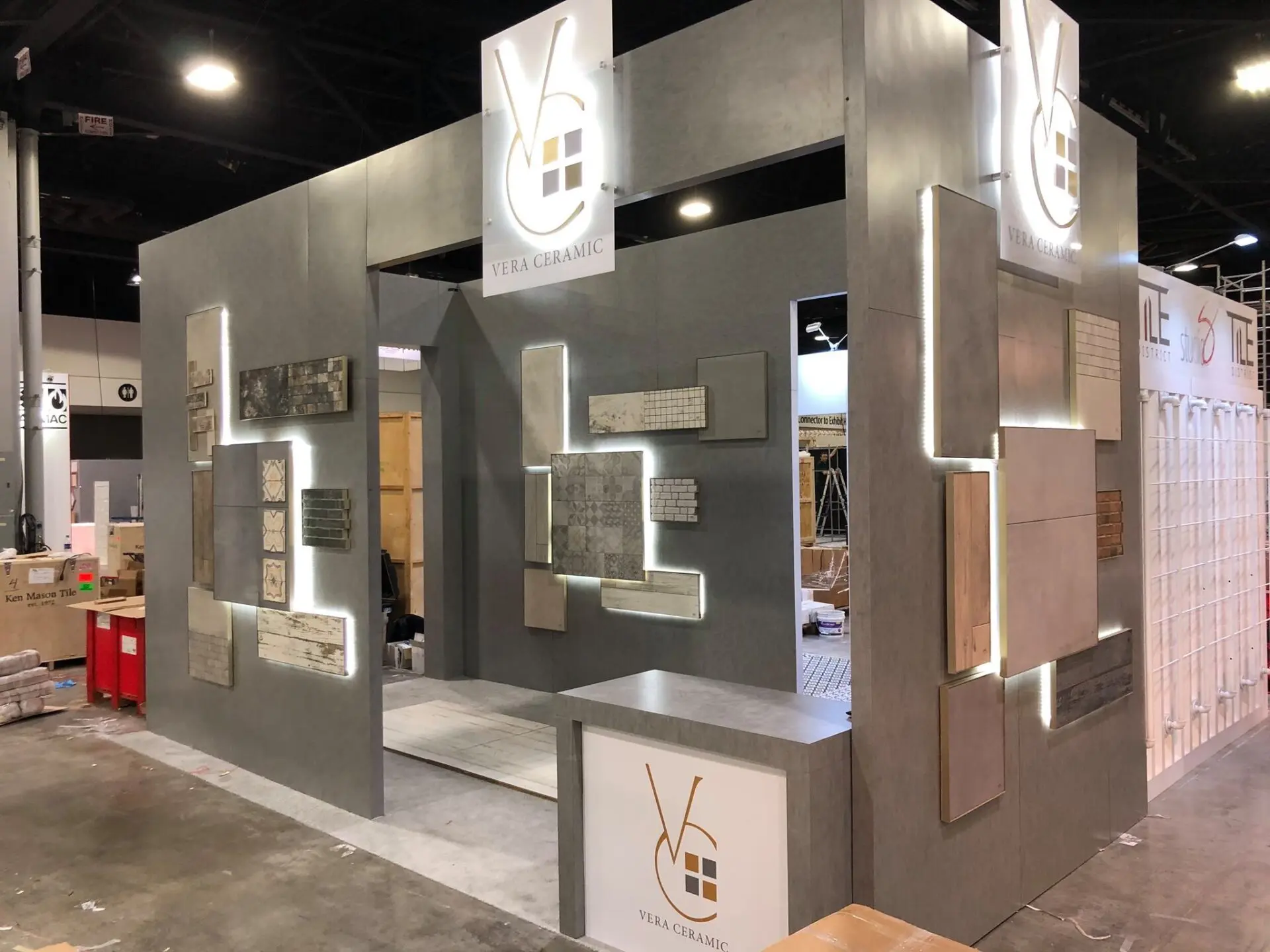 An exhibition booth for Vera Ceramic is displayed at a trade show. The booth features various tile samples arranged on walls with illuminated backlighting. The company's logo is prominently displayed at the top of two corners of the structure.